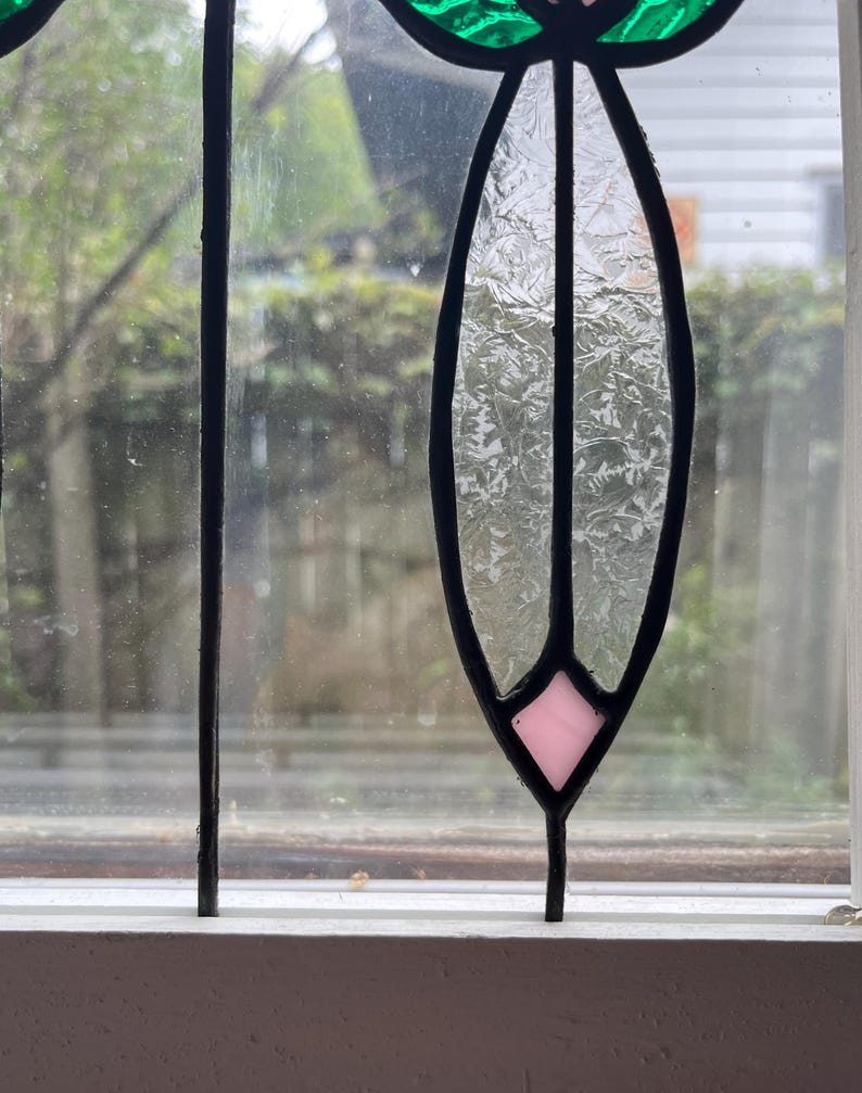 May include: A stained glass window panel featuring a vertical, elongated oval shape with textured, clear glass panes. The design includes a small, light pink diamond-shaped piece at the bottom and a vibrant green glass section at the top, all framed in black.
