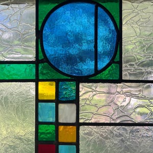 May include: A stained glass panel featuring a large central blue circle set against green glass. Below the circle is a grid of smaller square glass tiles in yellow, blue, white, green, and red. The surrounding panels consist of textured clear glass.