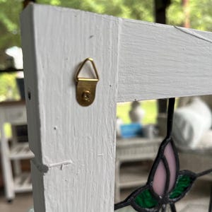 May include: A white painted wooden frame featuring a brass-colored triangular metal hanger attached to the side. A portion of a stained glass panel with pink and green glass pieces held together by dark metal solder lines is visible in the frame.