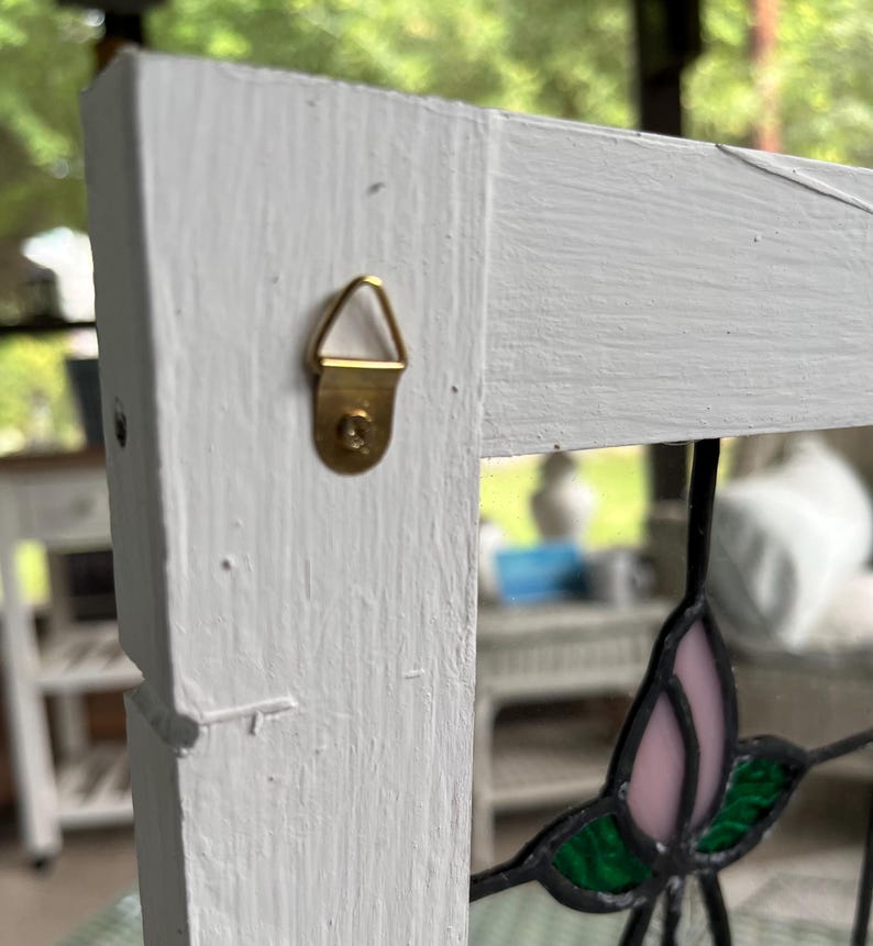 May include: A white painted wooden frame featuring a brass-colored triangular metal hanger attached to the side. A portion of a stained glass panel with pink and green glass pieces held together by dark metal solder lines is visible in the frame.
