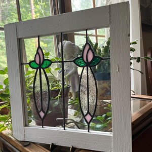 May include: A square white wooden window frame containing two stained glass panels. Each panel features a pink tulip bud at the top, green leaves, and a clear textured glass stem section, all held together by black metal lead lines.