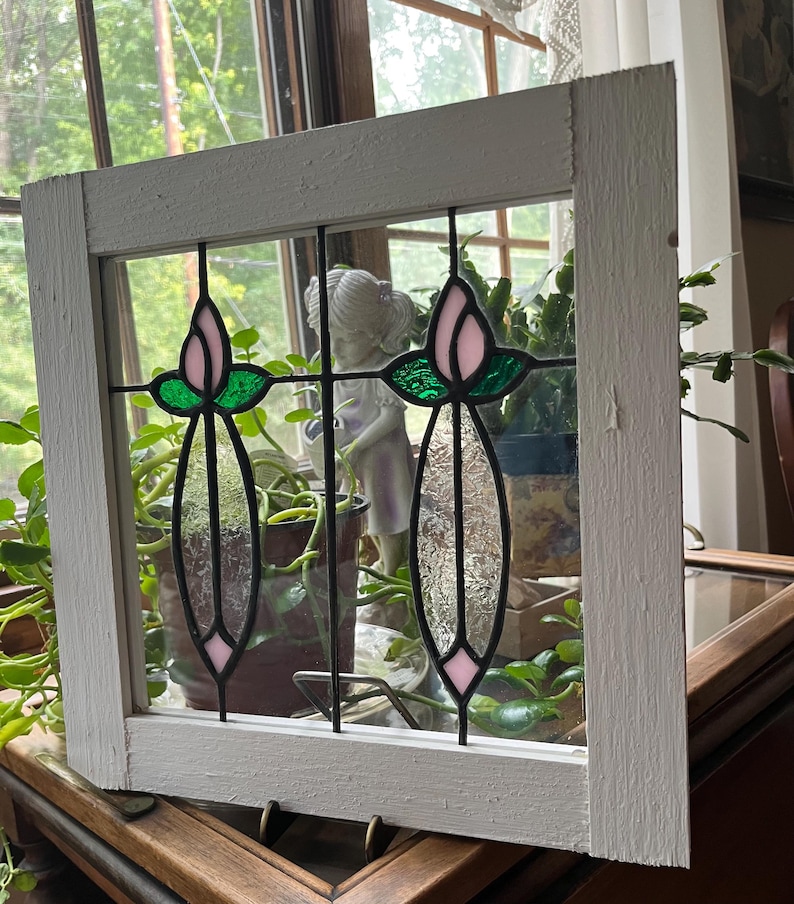 May include: A square white wooden window frame containing two stained glass panels. Each panel features a pink tulip bud at the top, green leaves, and a clear textured glass stem section, all held together by black metal lead lines.