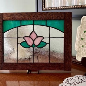 May include: A rectangular stained glass panel framed in dark brown wood. The glass features a central pink lotus flower design with green leaves, set against a clear, textured background with a horizontal band of green glass across the top.