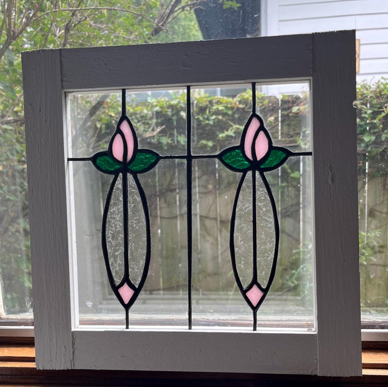 May include: A square window frame painted white containing a stained glass insert. The glass features two symmetrical floral motifs with pink tulip buds, green leaves, and clear textured glass stems set within black leaded lines.
