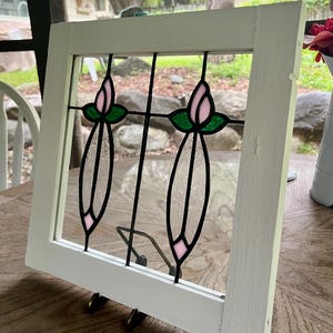May include: A square white wooden frame containing a stained glass panel. The glass features two vertical floral designs with pink tulip-shaped buds, green leaves, and clear textured glass stems, all held together by black metal lead lines.