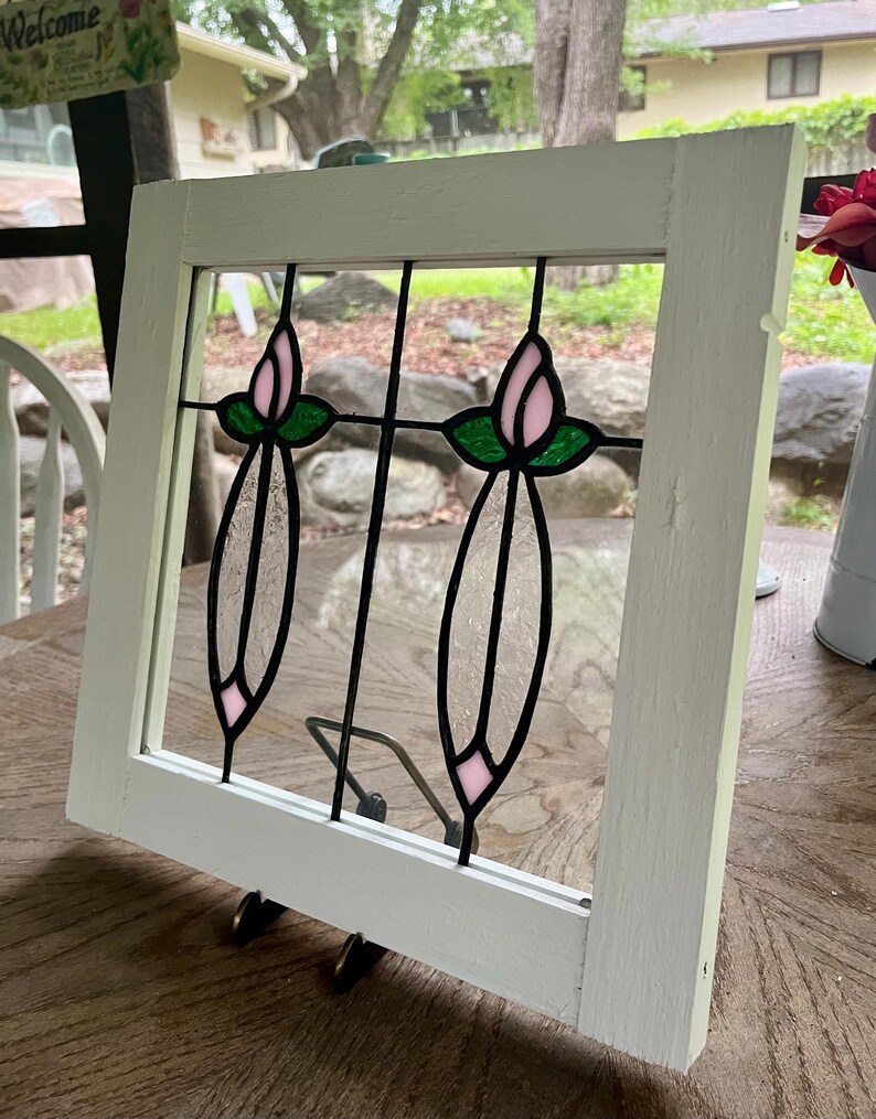 May include: A square white wooden frame containing a stained glass panel. The glass features two vertical floral designs with pink tulip-shaped buds, green leaves, and clear textured glass stems, all held together by black metal lead lines.