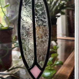 May include: A stained glass suncatcher featuring a vertical, leaf-shaped panel of textured, clear glass with a jagged, crystalline pattern. The piece is framed in black metal solder and includes a single, small, light pink diamond-shaped glass accent.