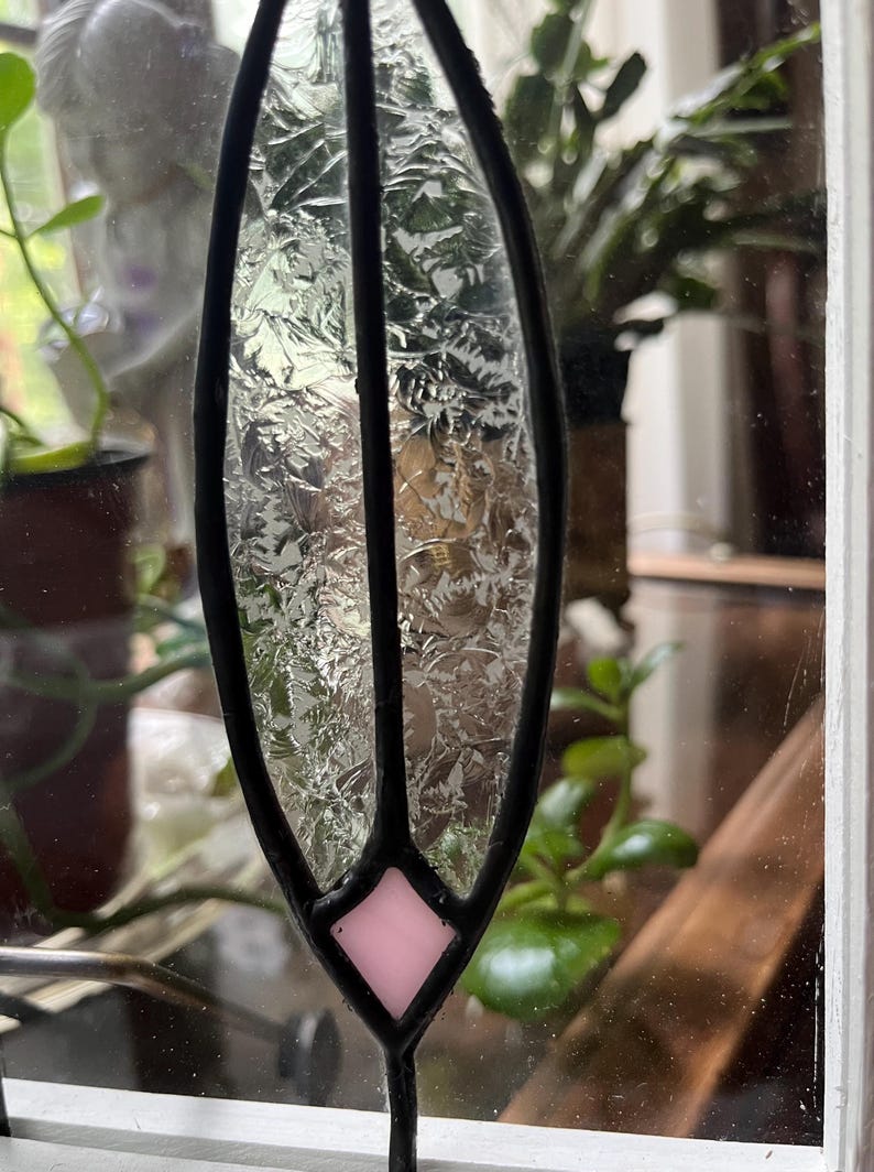 May include: A stained glass suncatcher featuring a vertical, leaf-shaped panel of textured, clear glass with a jagged, crystalline pattern. The piece is framed in black metal solder and includes a single, small, light pink diamond-shaped glass accent.