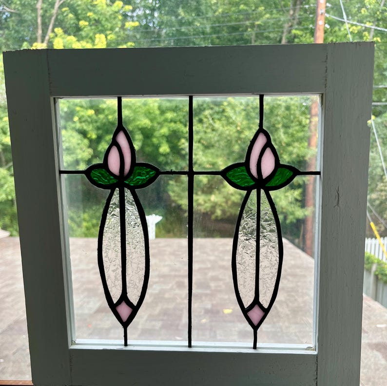 May include: A square white wooden window frame containing two stained glass panels. Each panel features a pink tulip bud with green leaves above a textured clear glass stem and a small pink diamond shape at the bottom, all joined by black lead.