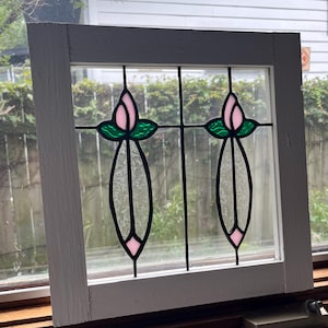 May include: A square white wooden window frame containing two stained glass panels. Each panel features a pink tulip bud with green leaves above a long, textured clear glass stem and a small pink diamond shape at the bottom base.