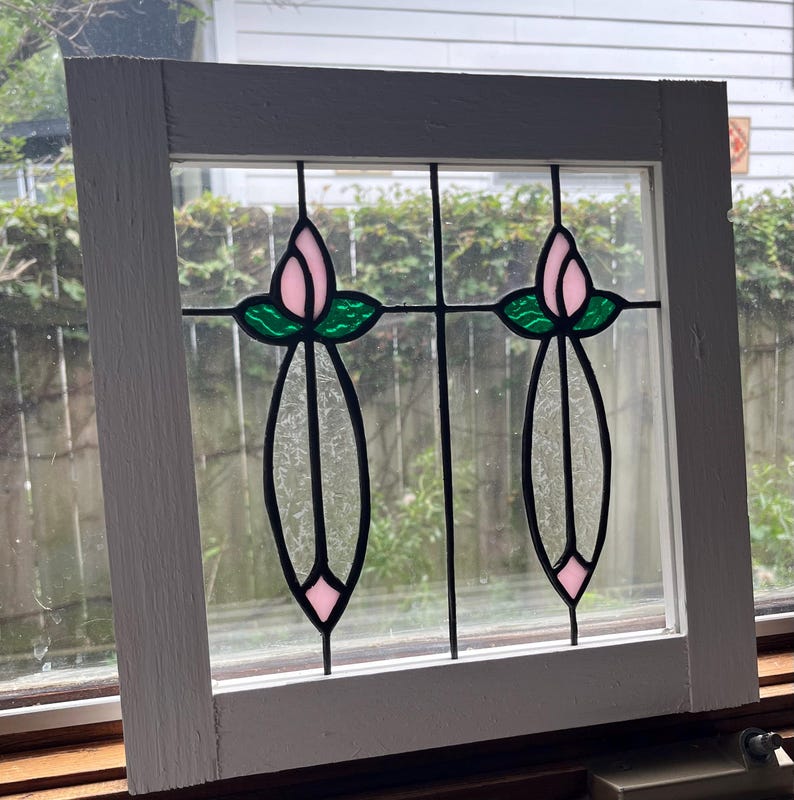 May include: A square white wooden window frame containing two stained glass panels. Each panel features a pink tulip bud with green leaves above a long, textured clear glass stem and a small pink diamond shape at the bottom base.