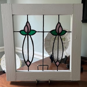 May include: A square white wooden frame containing two stained glass panels. Each panel features a pink tulip bud at the top, green leaves, and a clear textured glass stem section, all held together by black metal lead lines in a vertical design.