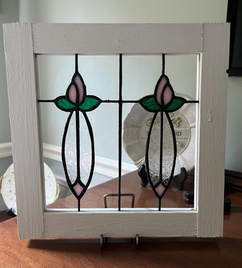 May include: A square white wooden frame containing two stained glass panels. Each panel features a pink tulip bud at the top, green leaves, and a clear textured glass stem section, all held together by black metal lead lines in a vertical design.