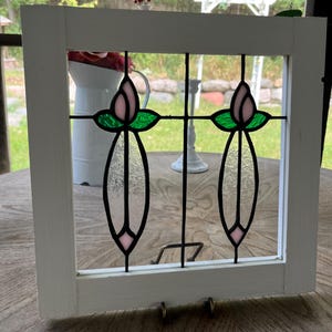 May include: A square white wooden frame containing a stained glass panel. The glass features two symmetrical floral motifs with pale pink buds, vibrant green leaves, and elongated clear textured glass stems, all joined by black metal lead lines.