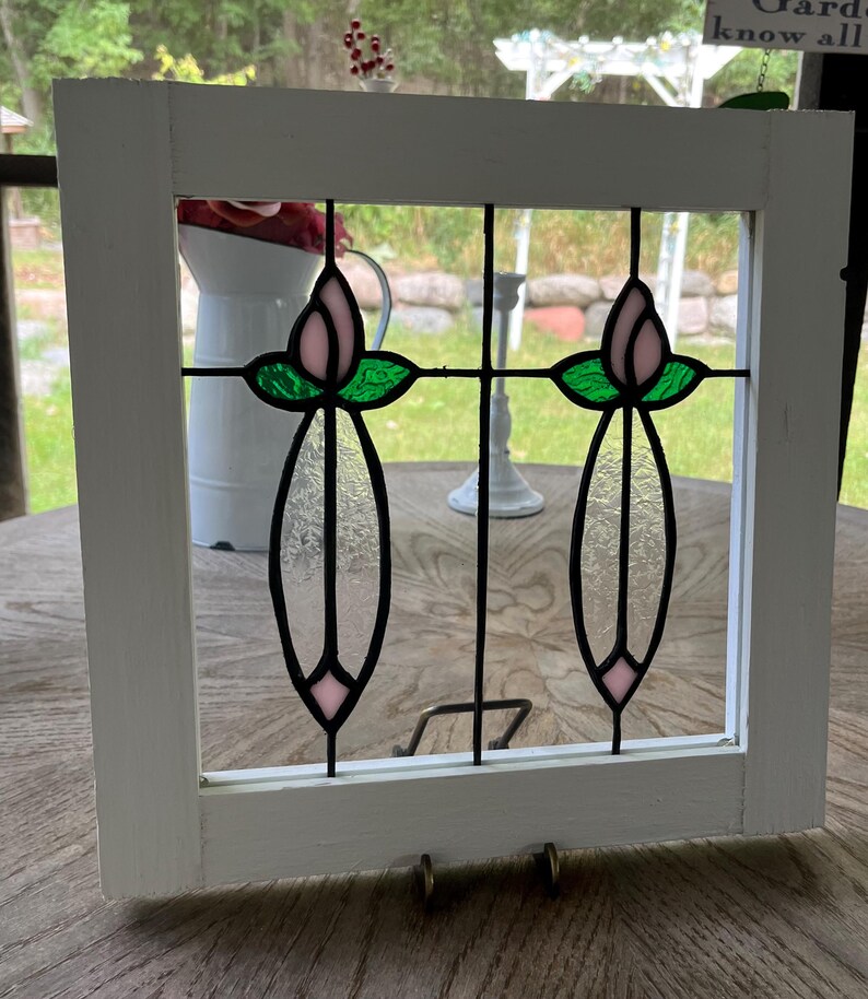 May include: A square white wooden frame containing a stained glass panel. The glass features two symmetrical floral motifs with pale pink buds, vibrant green leaves, and elongated clear textured glass stems, all joined by black metal lead lines.