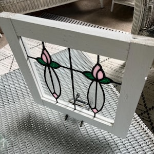 May include: A rectangular white wooden window frame containing a stained glass insert. The glass features two symmetrical floral motifs with pink petals, green leaves, and clear glass stems, all outlined in black metal cames within the frame.