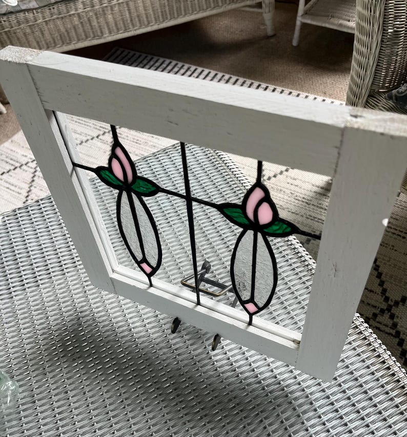 May include: A rectangular white wooden window frame containing a stained glass insert. The glass features two symmetrical floral motifs with pink petals, green leaves, and clear glass stems, all outlined in black metal cames within the frame.