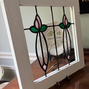 May include: A square white wooden frame containing a stained glass panel with two vertical floral motifs. Each motif features a pale pink bud, green leaves, and a textured clear glass stem held together by black metal cames within the frame.
