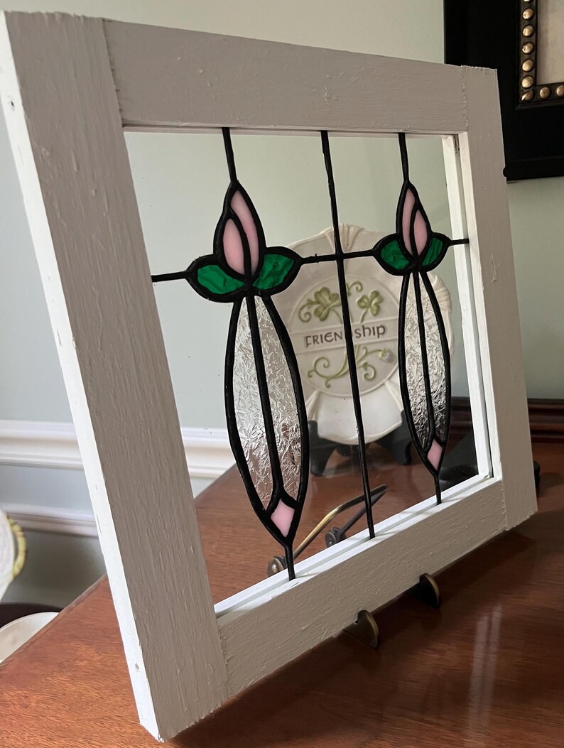 May include: A square white wooden frame containing a stained glass panel with two vertical floral motifs. Each motif features a pale pink bud, green leaves, and a textured clear glass stem held together by black metal cames within the frame.