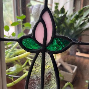 May include: A stained glass panel featuring a central floral motif with a pale pink bud, two textured emerald green leaves, and a clear, patterned glass stem. The individual glass pieces are joined by dark, soldered lead lines.