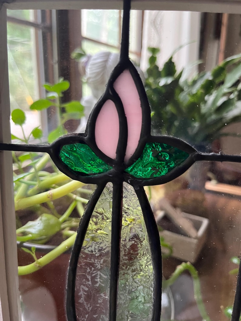 May include: A stained glass panel featuring a central floral motif with a pale pink bud, two textured emerald green leaves, and a clear, patterned glass stem. The individual glass pieces are joined by dark, soldered lead lines.