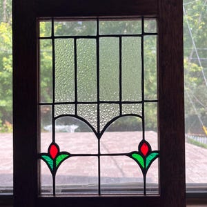 May include: A rectangular stained glass window panel framed in dark wood. The design features clear textured glass panes in the upper section and two stylized red and green floral motifs at the bottom, all held together by dark metal lead lines.