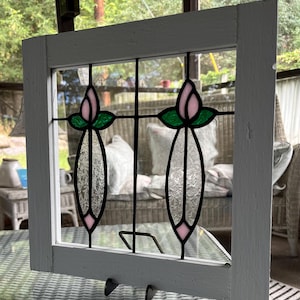 May include: A square white wooden frame containing a stained glass panel. The panel features two vertical floral designs with pink glass petals, green glass leaves, and textured clear glass stems, all held together by black metal lead lines.