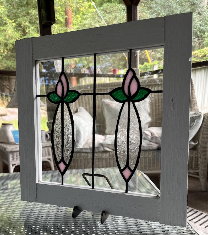 May include: A square white wooden frame containing a stained glass panel. The panel features two vertical floral designs with pink glass petals, green glass leaves, and textured clear glass stems, all held together by black metal lead lines.