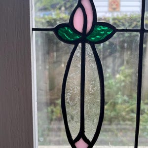 May include: A stained glass window panel featuring a central vertical design with a light pink bud at the top, two vibrant green leaves, and a long, textured, clear glass stem that tapers to a small light pink diamond shape at the bottom.