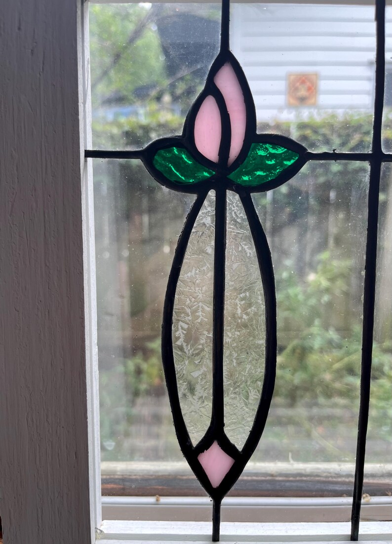 May include: A stained glass window panel featuring a central vertical design with a light pink bud at the top, two vibrant green leaves, and a long, textured, clear glass stem that tapers to a small light pink diamond shape at the bottom.