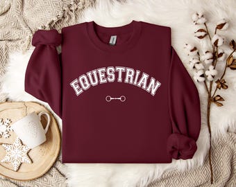 May include: A maroon long-sleeve crewneck sweatshirt featuring the word EQUESTRIAN in white block letters with a thin white outline, arched over a small white graphic of a horse bit. The garment has a ribbed collar, cuffs, and hem.