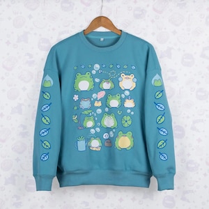 Könnte beinhalten: Türkisfarbenes Sweatshirt mit einem Muster aus Cartoon-Fröschen in verschiedenen Posen. Die Frösche sind hauptsächlich grün und weiß, mit gelben Akzenten. Die Ärmel haben Blatt- und Froschmotive. Das Sweatshirt hängt an einem Holzkleiderbügel.