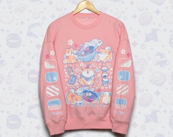 May include: A pink crewneck sweatshirt featuring a colorful graphic of Shiba Inu dogs, ramen bowls, and other Japanese-themed illustrations. The sleeves have additional decorative elements. The sweatshirt is hanging on a wooden hanger.