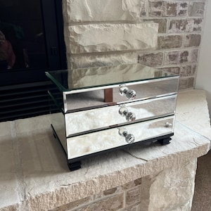 May include: A mirrored jewelry box with three drawers and crystal-like knobs. The box is rectangular with a glass top and sits on a black base. The background features a brick fireplace.