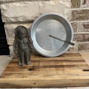 May include: A vintage metal pie pan with the text "Bake King" and a small metal angel statue are displayed on a wooden cutting board. The pie pan is silver and has a handle. The angel statue is gray and has wings.