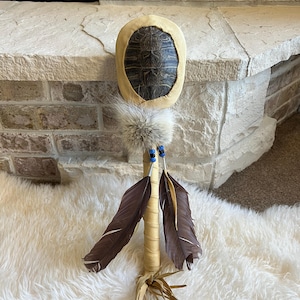 May include: A handcrafted Native American rattle featuring a turtle shell, adorned with a tan leather frame, fluffy fur, blue beads, and brown feathers. The handle is wrapped in tan material, with a fringe of tan leather at the base.