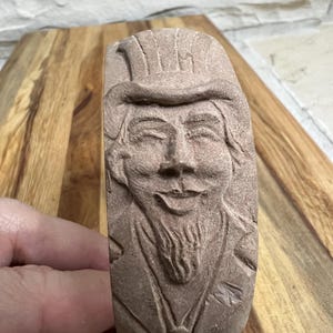 May include: A brown clay sculpture of a person's face with a hat. The sculpture is carved in detail and has a whimsical expression.
