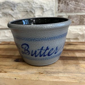 May include: A gray stoneware butter crock with a dark interior and blue stripes. The word "Butter" is written in blue script on the side. The crock is sitting on a wooden surface.