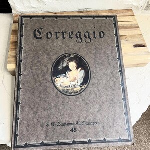 May include: A brown book with a black border and the word "Correggio" in black lettering on the front cover. The book features a painting of a woman holding a baby with a donkey in the background. The book also has the text "G. H. Seemanns Künstlermappen 45" on the bottom.
