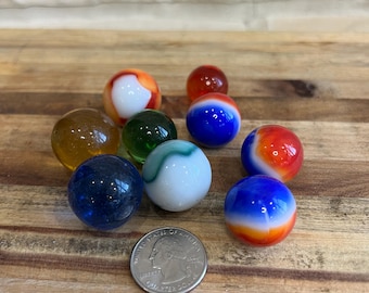 May include: A collection of ten glass marbles in various colors and patterns, including red, white, blue, green, and yellow. The marbles are arranged on a wooden surface next to a US quarter.