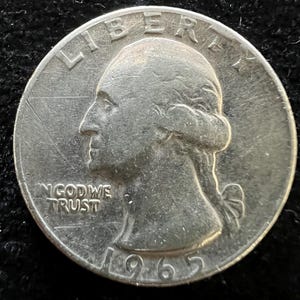 May include: A silver 1965 quarter coin. The obverse features a profile of a person with the inscriptions "LIBERTY" and "IN GOD WE TRUST". The year "1965" is visible at the bottom of the coin.