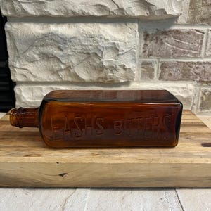 May include: A rectangular amber glass bottle with the embossed text "LASH'S BITTERS" is displayed horizontally. The bottle is positioned on a wooden cutting board, with a brick wall in the background. The bottle has a long neck and a flat, rectangular body.