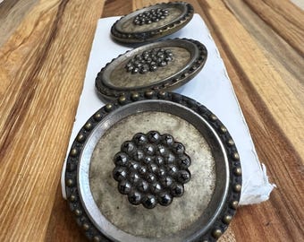 May include: Three antique silver and brass drawer pulls with a raised, circular design of small, black metal balls. The pulls are arranged in a row on a wooden surface.
