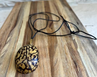 May include: A brown and black patterned seed pod pendant on a black cord necklace. The pendant is oval shaped and has a natural wood look.