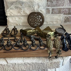 May include: A collection of antique brass and black metal drawer pulls and cabinet hardware. The hardware includes a variety of styles, including ornate, simple, and geometric. The hardware is displayed on a wooden surface.