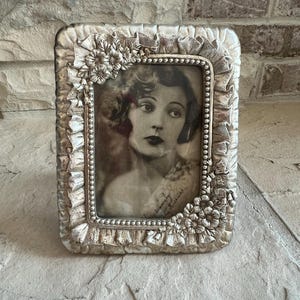 May include: Silver-toned rectangular picture frame with a vintage black and white photograph of a woman. The frame has a textured, ornate design with floral and beaded accents. The photograph is signed in cursive.