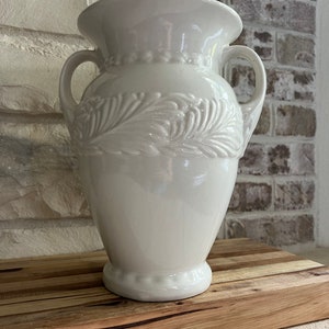 May include: A white ceramic vase with two handles and a decorative embossed leaf design. The vase has a wide opening and a pedestal base. The vase is sitting on a wooden surface.