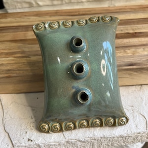 May include: A rectangular ceramic flower frog with a glossy sage green glaze. The piece features three raised circular openings in the center for stems and decorative spiral scrollwork along the top and bottom edges in a muted tan color.