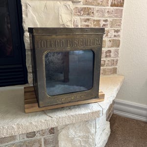 May include: A vintage rectangular metal Toledo Biscuit Co. storage tin with a dark brass-toned finish. The front features a large glass viewing window framed by a beaded border, with the text TOLEDO BISCUIT CO. embossed at the top and TOLEDO at the bottom.