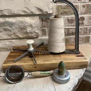 May include: A collection of vintage kitchen tools displayed on a wooden cutting board. Includes a metal and wood flour sifter, a biscuit cutter, a twine holder, and a pie cutter. The tools are arranged on a light brown wooden surface.